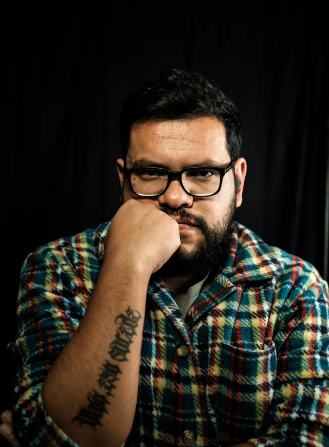 Retrato de Abraham Reyes con camisa a cuadros, la mano en el mentón, sobre fondo negro